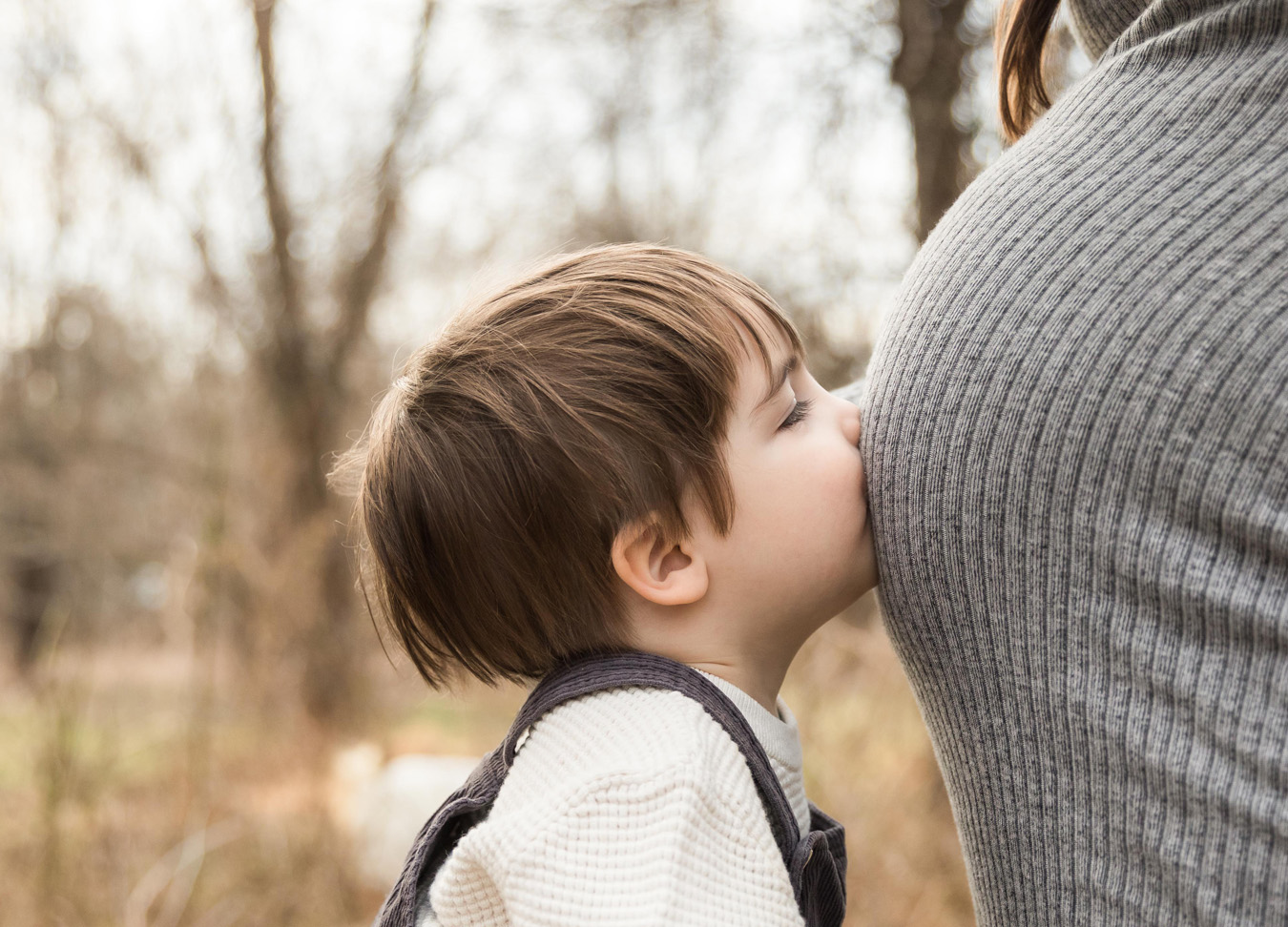 Best doulas in Northern Virginia series featuring a toddler with brown hair, a white sweatshirt and brown overalls kissing his mother's baby belly