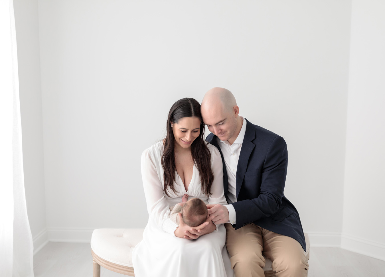A mom in a white dress and a dad in a navy blazer and khaki pants sitting on a bench holding their baby boy during a studio newborn session