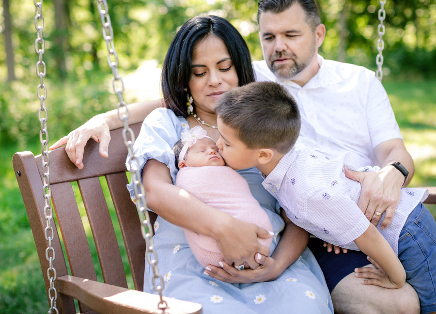 newborn photos in D.C. featuring a family of four sitting on a bench while the big brother kisses his little sister