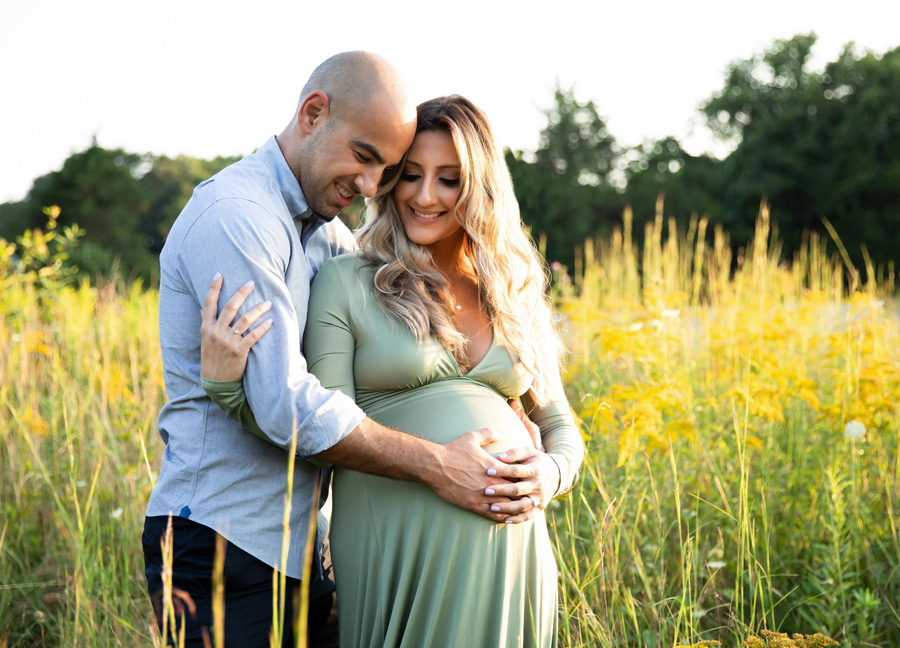 Husband hugging his pregnant wife while standing in a field at sunset.