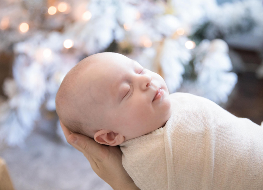 A mother holding her swaddled baby boy in front of a Christmas tree.