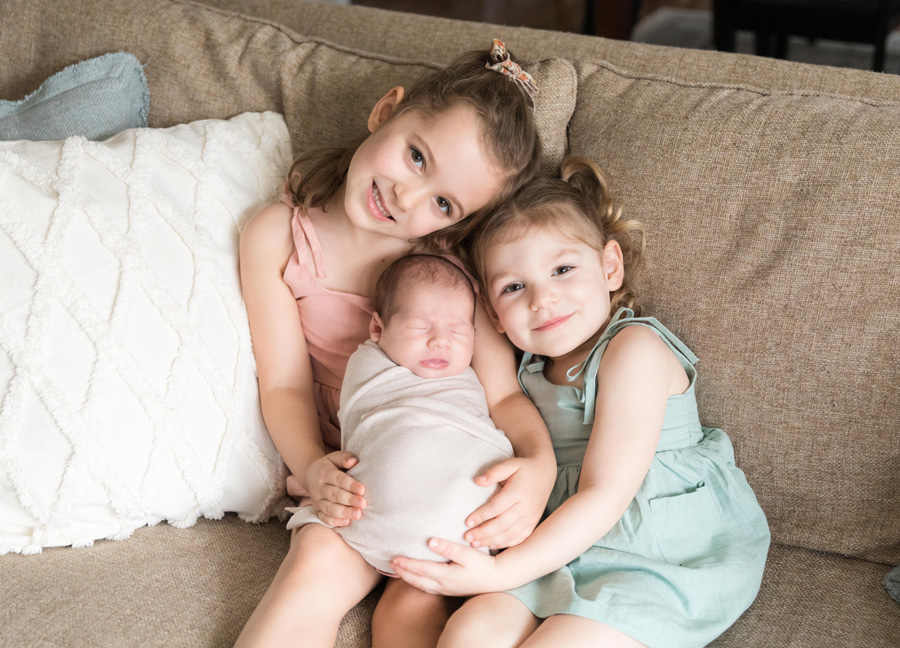 newborn photography with siblings featuring two sisters holding their baby sister on a couch