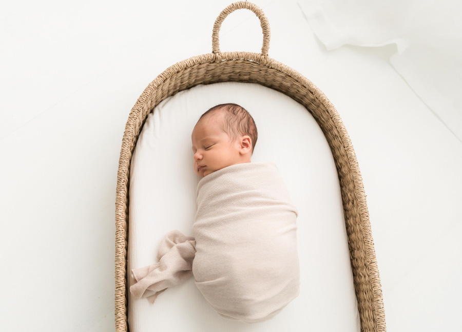 A baby boy with a cream swaddle sleeping in a moses basket.