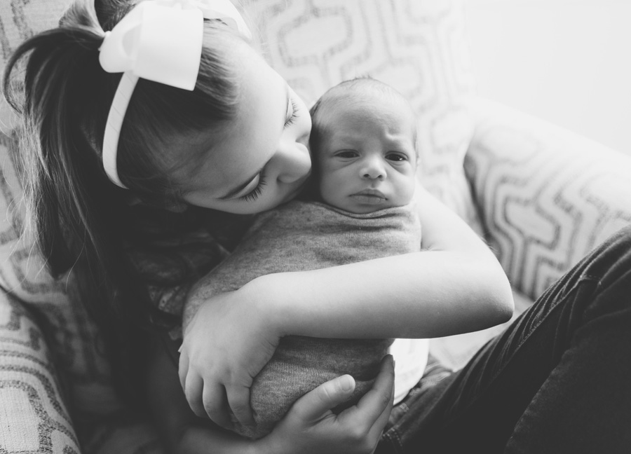 A black and white image of a girl kissing her newborn brother on the cheeks. 