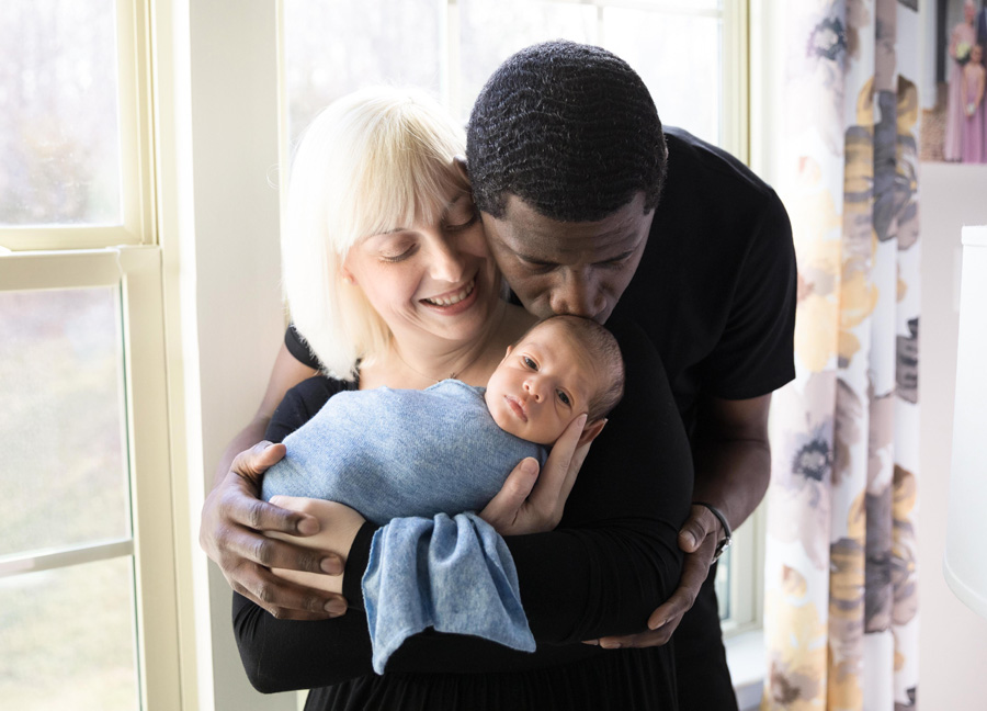New parents holding their newborn baby by the window in their living room during an at home newborn photography session in Northern Virginia.