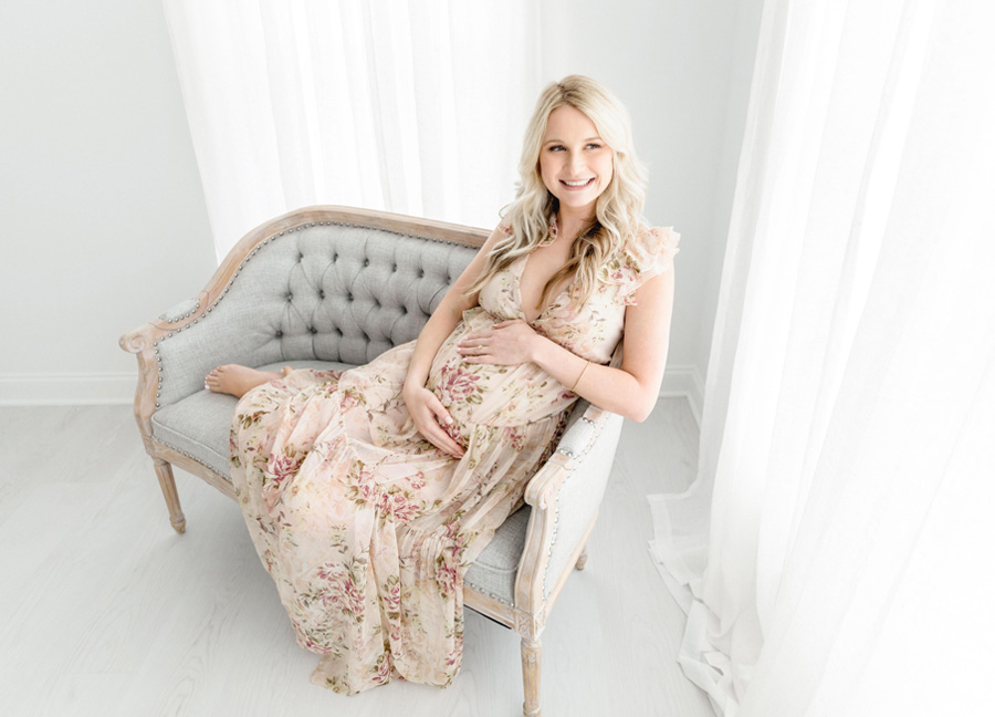 maternity photoshoot outfit featuring a mom sitting ona. couch smiling and holding her belly