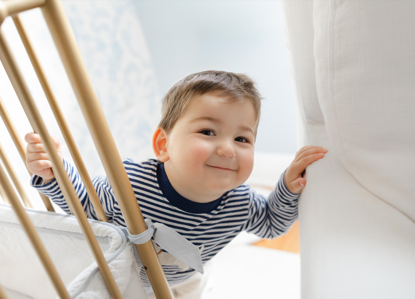 A toddler smiling into the camera while hiding behind the crib and rocking chair. 