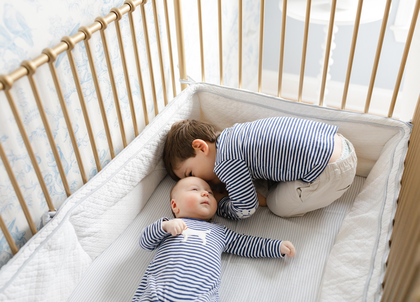 A toddler kissing his baby brother on the head while laying in the crib together. 