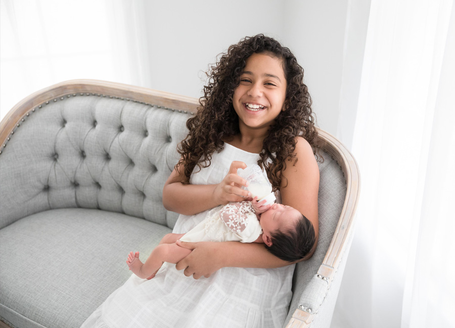 A girl laughing while bottle feeding her baby sister during their newborn photography session in Northern Virginia.
