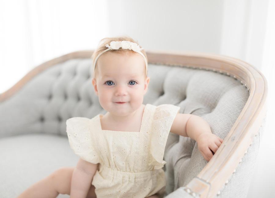 A baby girl in a beige onesie and floral headband smiling into the camera during her one year milestone session in Northern Virginia.