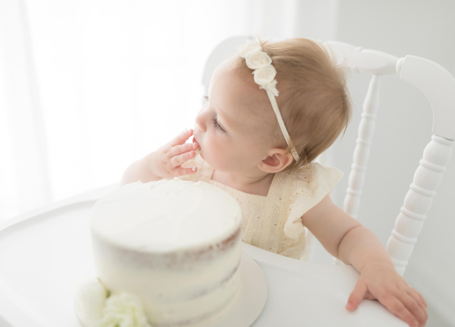 A baby girl in a beige romper and floral headband eating a naked cake during her cake smash session in Northern Virginia. 