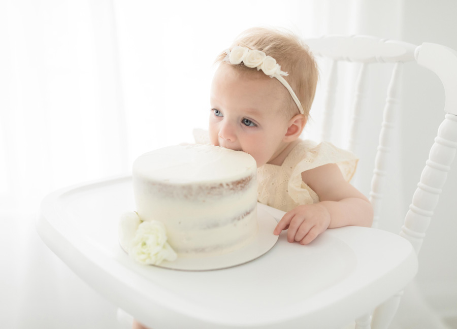 A baby girl in a beige romper and floral headband taking a bite out of a cake during her cake smash session in Northern Virginia. 