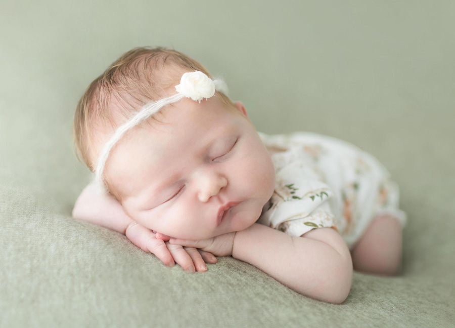 A baby girl in a floral onesie sleeping with her hands under her chin during her newborn session with Stephanie Honikel Photography. 