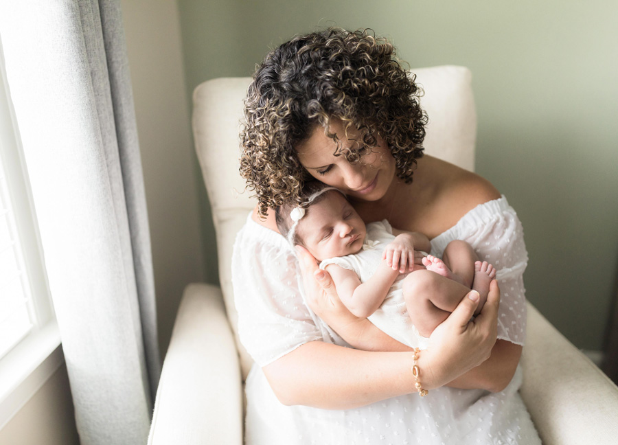 A mother in a white dress sitting in a rocking chair while holding her new baby girl during their DC newborn photography session.