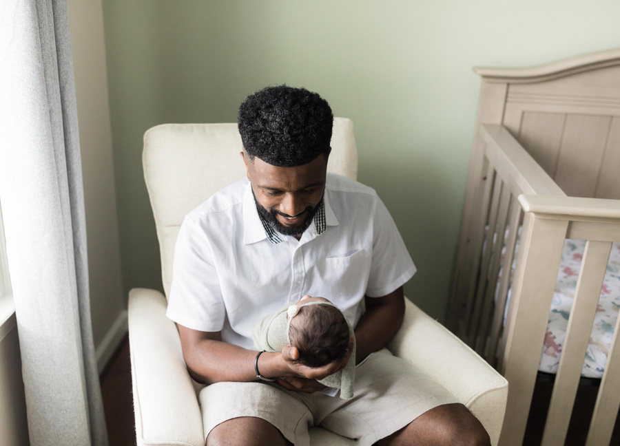 A father holding his swaddled baby girl while sitting in the rocking chair during their DC newborn photography session at home. 