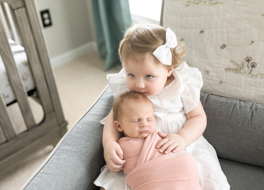 financially prepare for a newborn with newborn photography in Washington D.C.. This picture shows an older sister and younger sister together on the nursery chair.