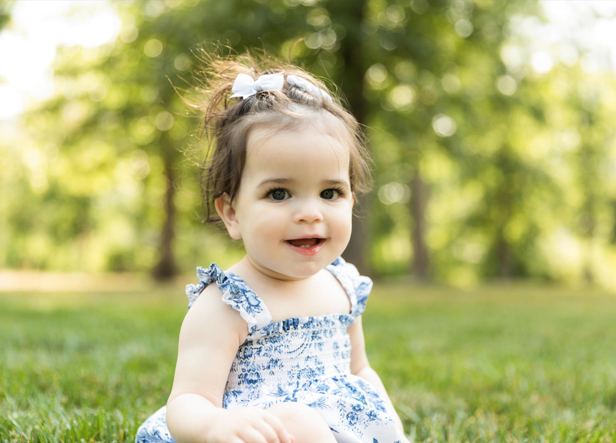 Northern virginia newborn photographer takes a picture of a little girl sitting in the grass smiling wearing a bow and a blue and white dress.