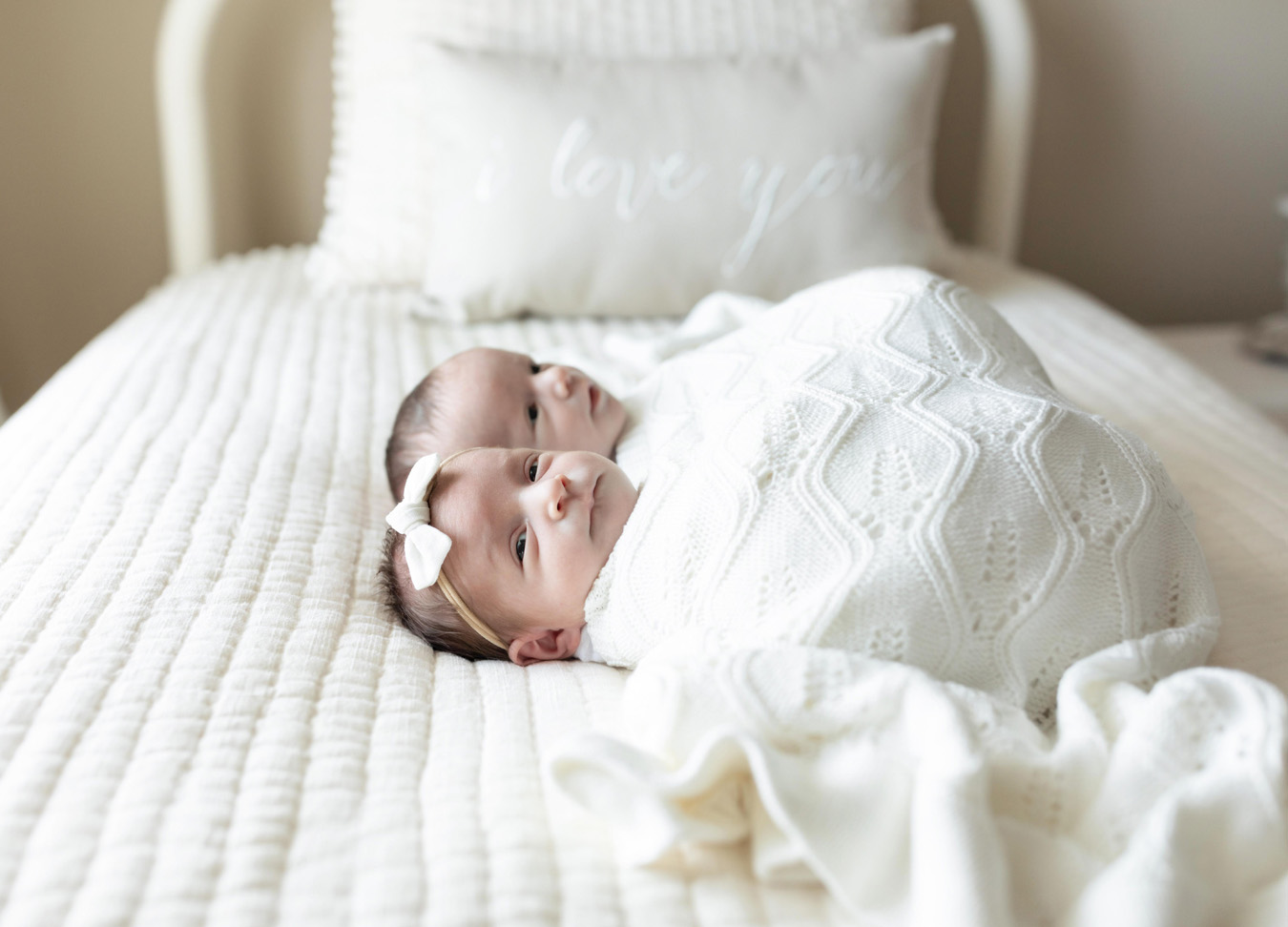 twins laying next to each other covered in a white blanket laying on a white bed for a twin newborn session