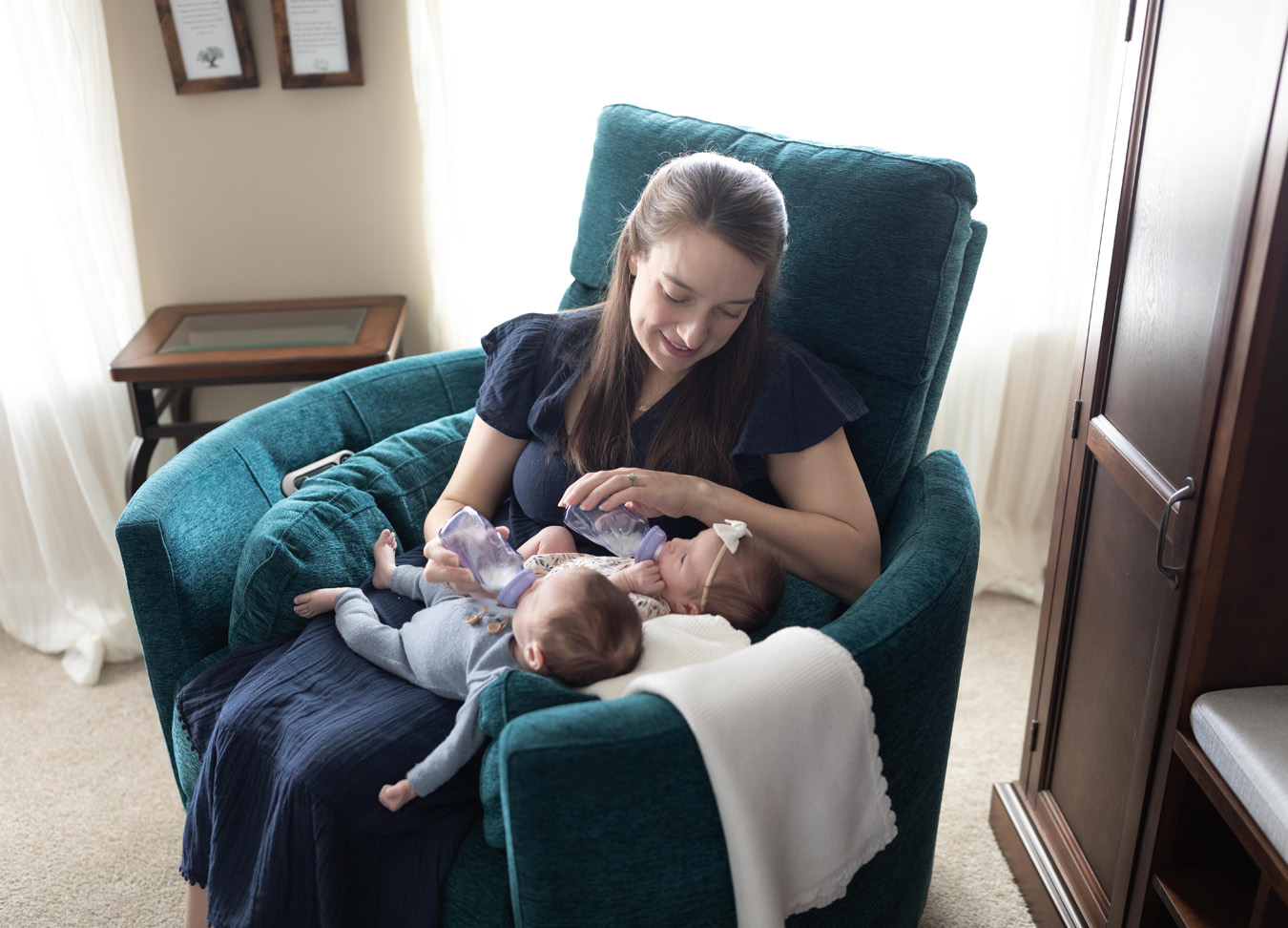 Mom feeding bottles to both twins while sitting in a green chair during a twin newborn session