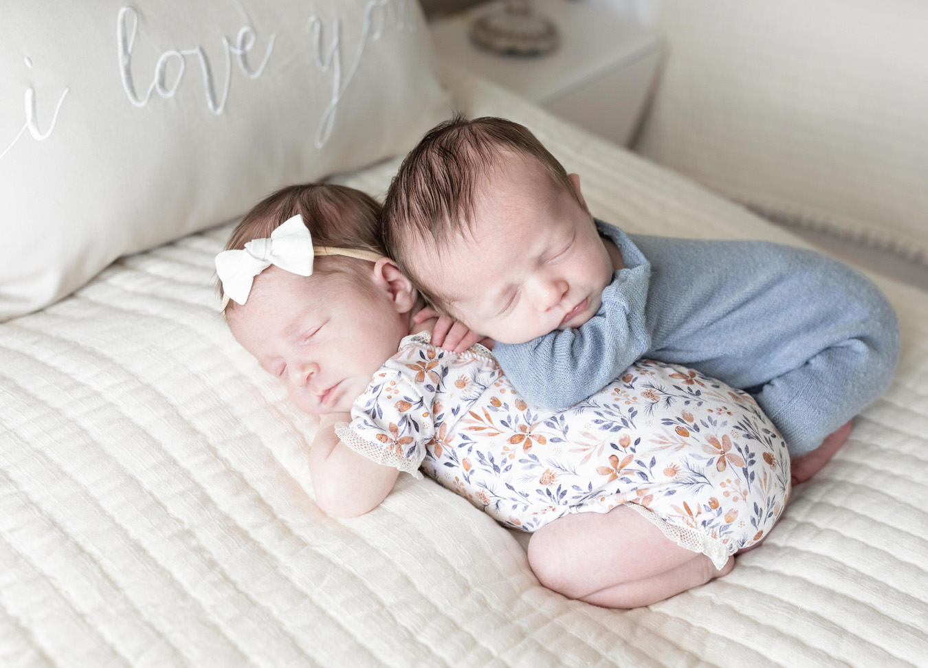twin newborn session with one twin laying on the bed in a floral outfit and the second twin leaning on top of the first twin in a blue outfit.