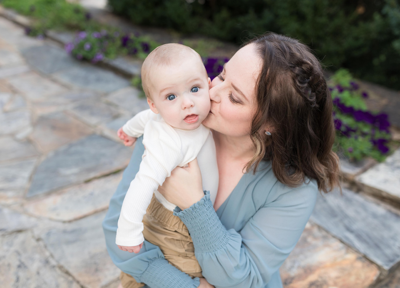 A blue-eyed baby boy looking into the camera while being kissed on the cheeks by his mom during a family session at Rust Manor in Leesburg