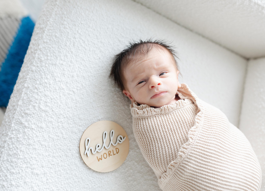 newborn baby wrapped up in a cozy blanket laying on the couch with a sign that says "hello world".