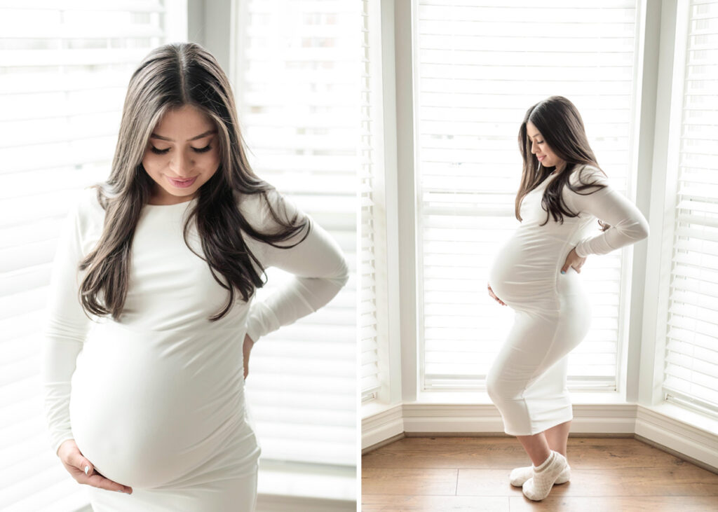 A mom to be in a white dress in a bay window, holding her baby belly