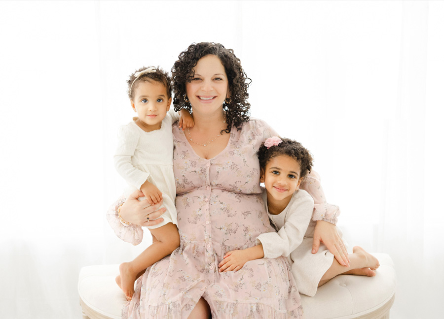 A mother to be in a pink blush floral dress sitting on a bench with her two small children during their simple studio maternity session 