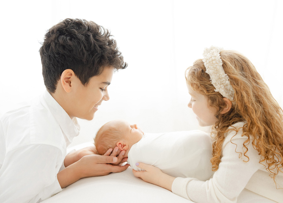 An older brother and sister holding at their newborn baby brother who is wrapped in a white swaddle