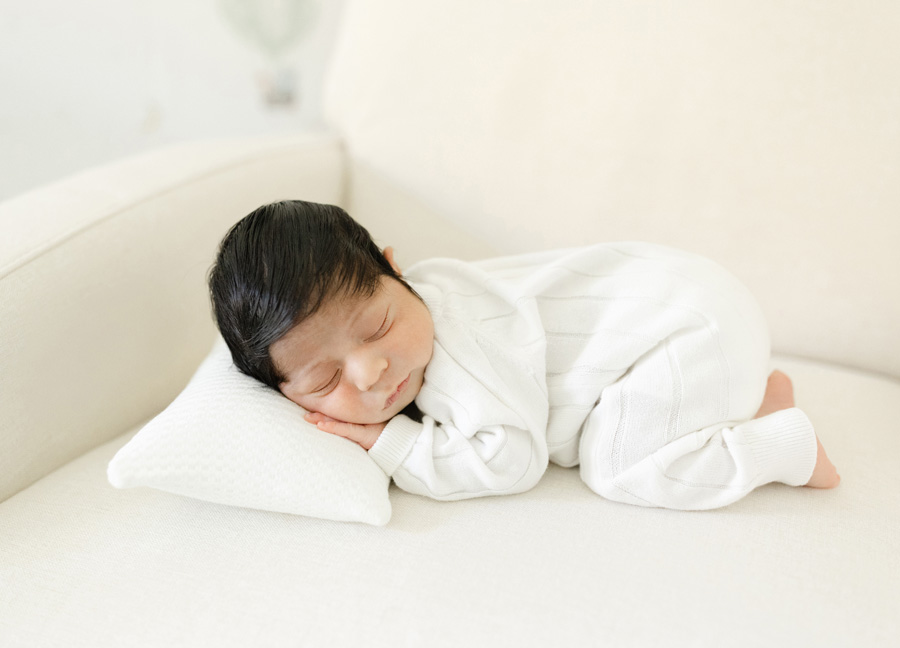 Newborn in a white outfit peacefully sleeping on a neutral rocking chair in a softly lit nursery.