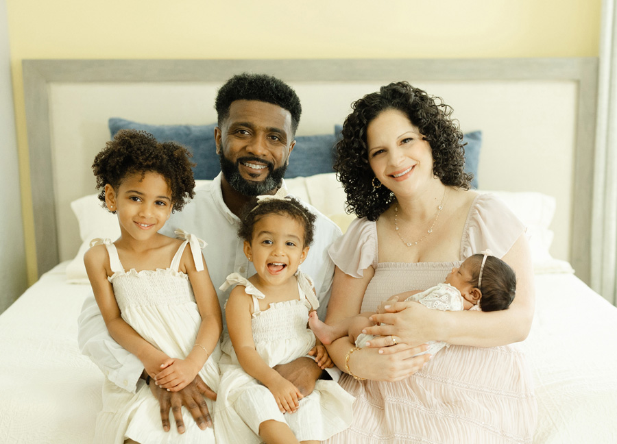 A joyful family of five, with two young girls in white dresses and a newborn, smiling together