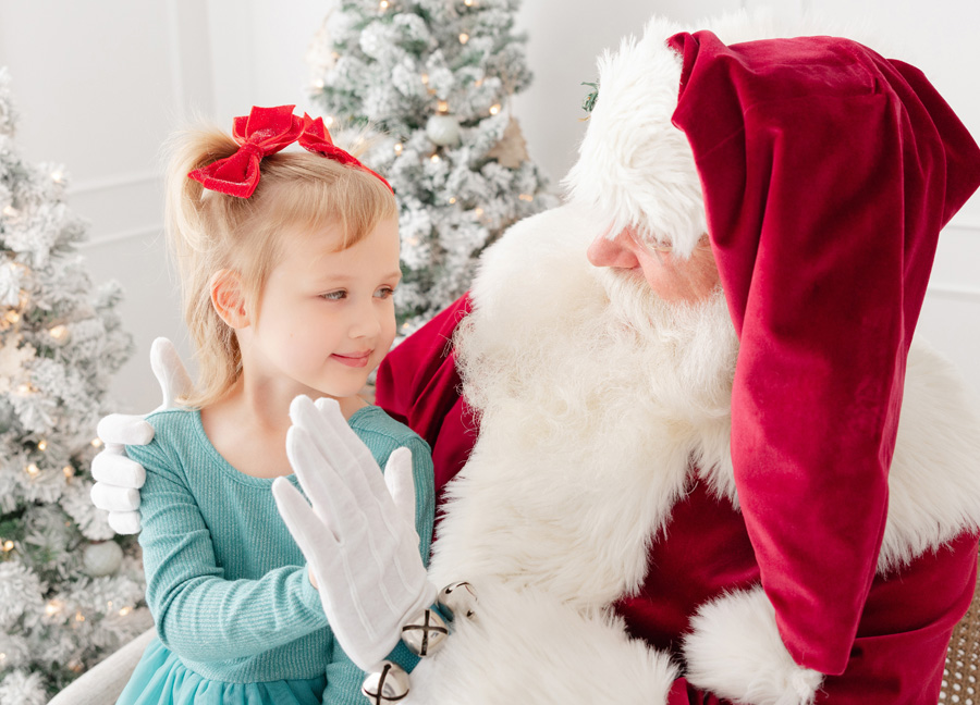 2025 Fun DC Holiday events for families: Young girl in blue dress with red bows meeting Santa during the Santa Photos Experience hosted by Stephanie Honikel Photography