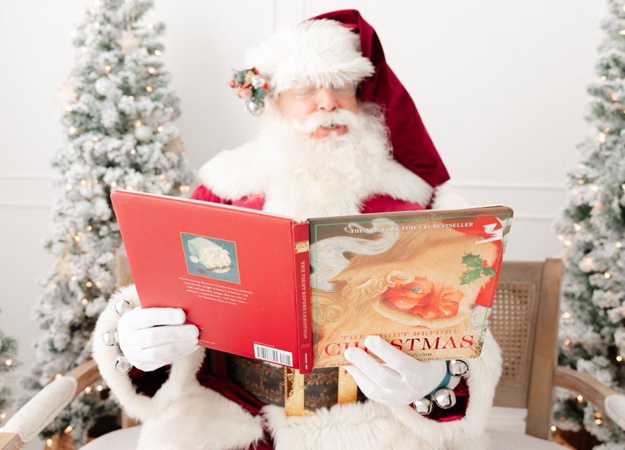 Santa Claus sitting on a brown bench surrounded by snowy Christmas trees reading the Christmas story 