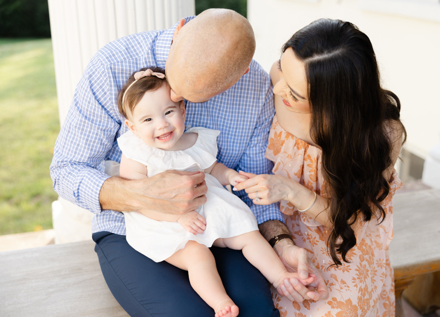 A family of three sitting on a bench with the baby girl in her dad's lap while he gently kisses her head