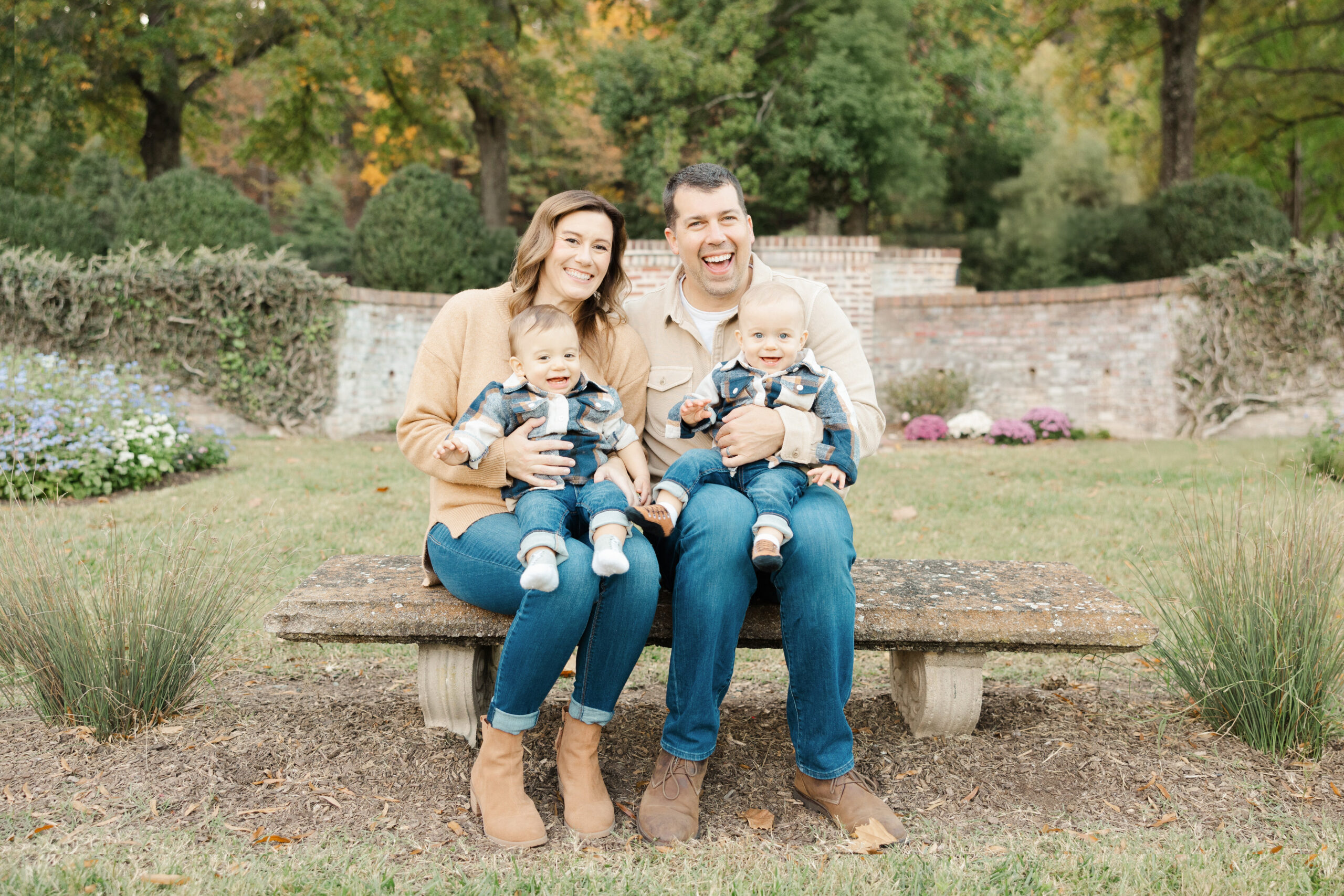 A smiling family of four sitting on a stone bench outdoors with their twin toddlers during a fall session. Family Photography in Northern Virginia by Honikel Photography.