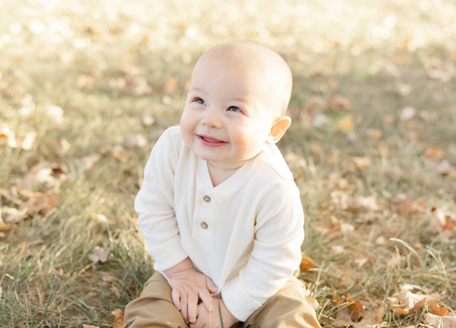 Smiling baby sitting on the grass during an outdoor fall session, dressed in a neutral outfit and surrounded by soft natural light and scattered autumn leaves.