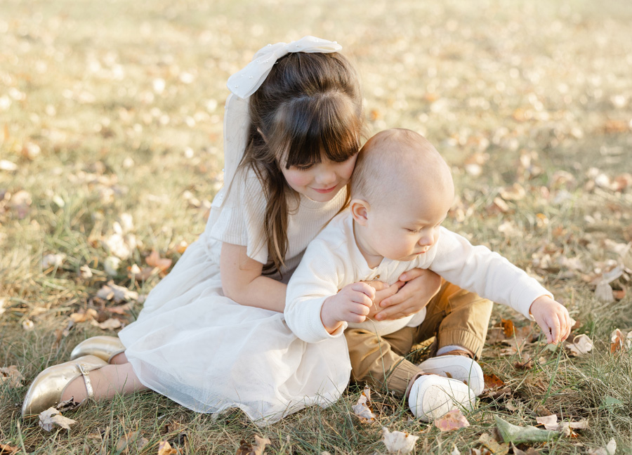 Older sister gently hugging her baby sibling during an outdoor fall photo session, sitting on the grass in soft natural light with scattered autumn leaves.