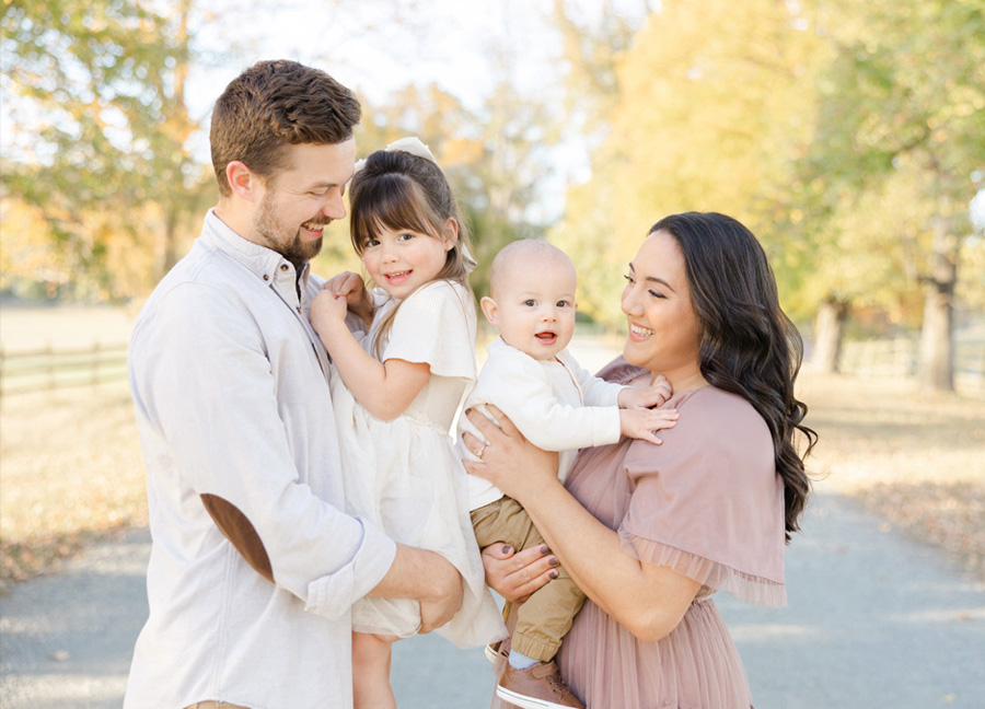 Parents holding their two young children during an outdoor fall family session, sharing smiles and connection along a tree-lined path with soft golden autumn light.