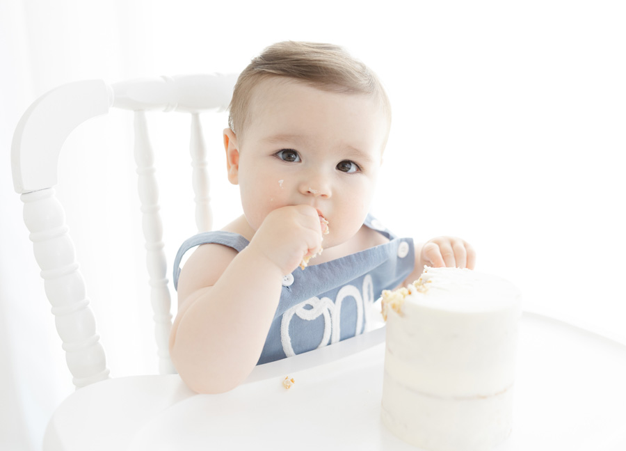 One-year-old baby boy in a blue “One” romper enjoys his first birthday cake smash during a Northern Virginia milestone photography session. He sits in a white high chair tasting frosting from a simple white birthday cake.