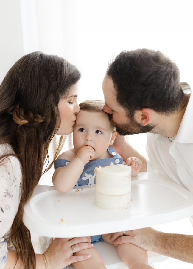 One-year-old baby boy enjoys his birthday cake while his parents kiss him on the head during a Northern Virginia cake smash photography session. This sweet family portrait captures a loving moment during his first birthday celebration.