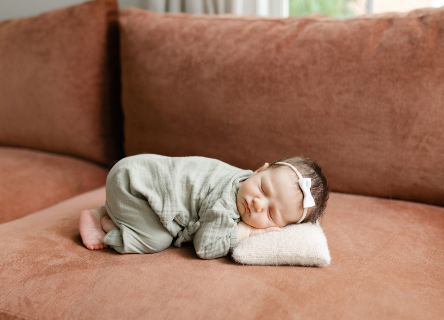 Newborn baby girl wearing a sage green outfit and delicate white bow, curled up asleep with her head resting on a soft cream pillow on a warm rust-colored sofa.