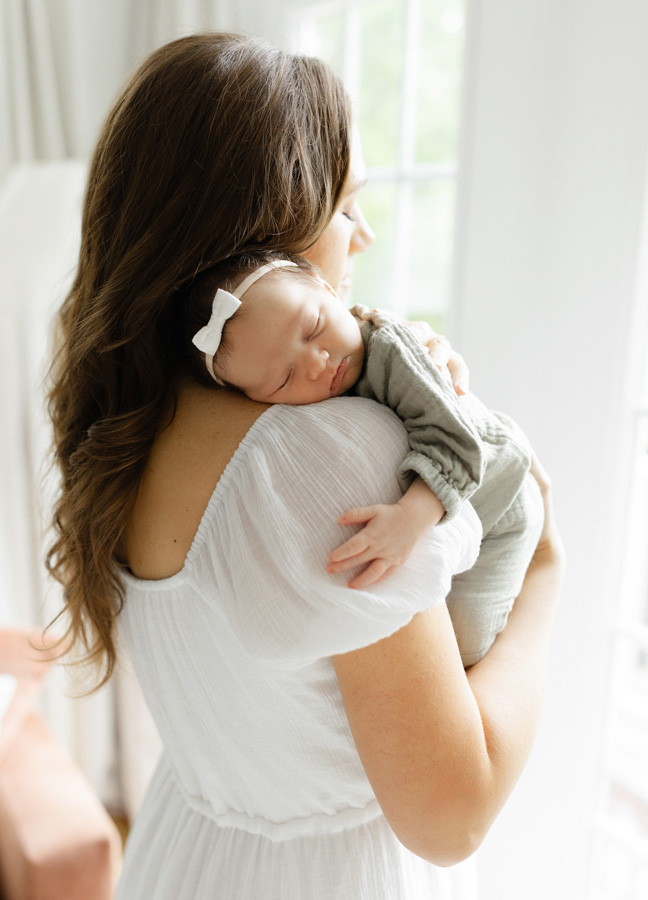 Mother in a flowing white dress holding her sleeping newborn baby girl over her shoulder, with baby resting peacefully in a sage green outfit and white bow beside a bright window.