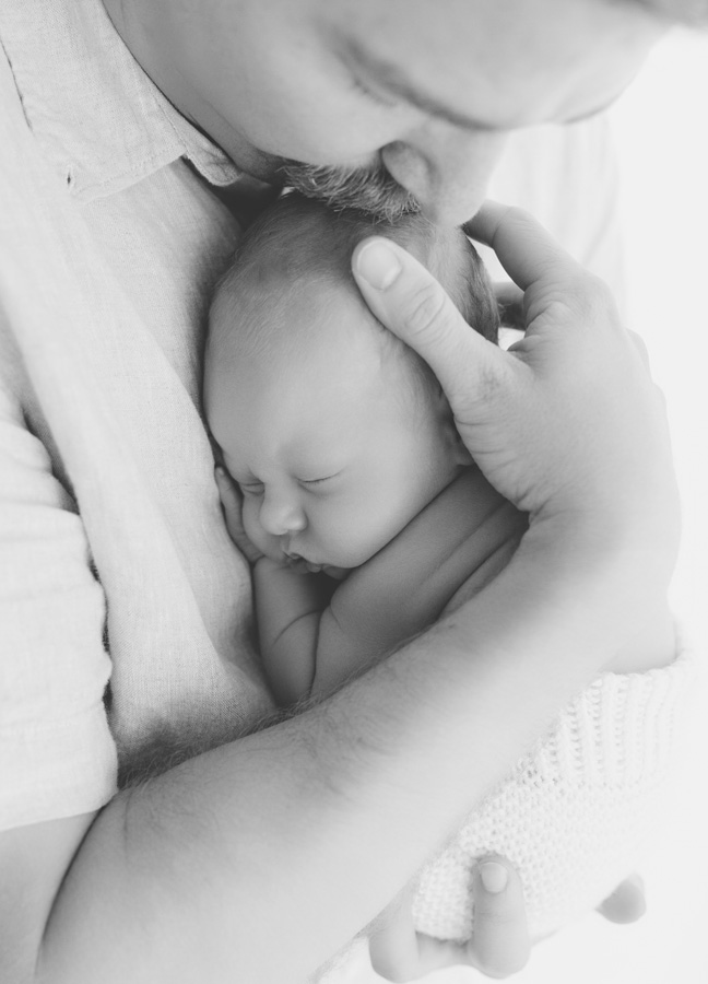 Black and white portrait of a father gently kissing his sleeping newborn baby while holding him close to his chest.