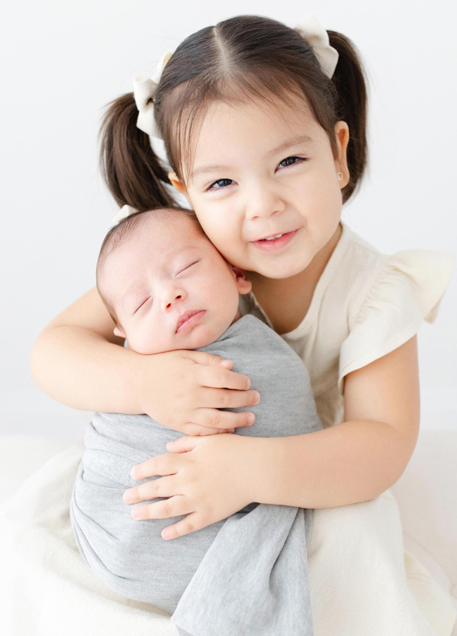 Smiling big sister gently hugging her sleeping newborn sibling during a bright studio portrait, a sweet example of newborn photos ideas Northern Virginia.