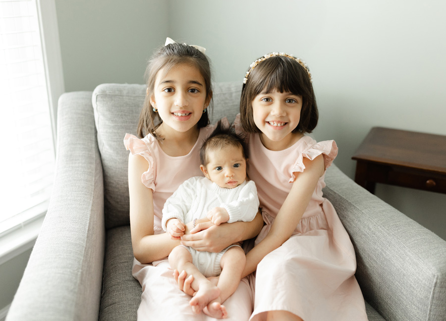 Two smiling big sisters cuddling their baby sibling on a cozy chair, a sweet family moment featuring newborn photos ideas Northern Virginia.