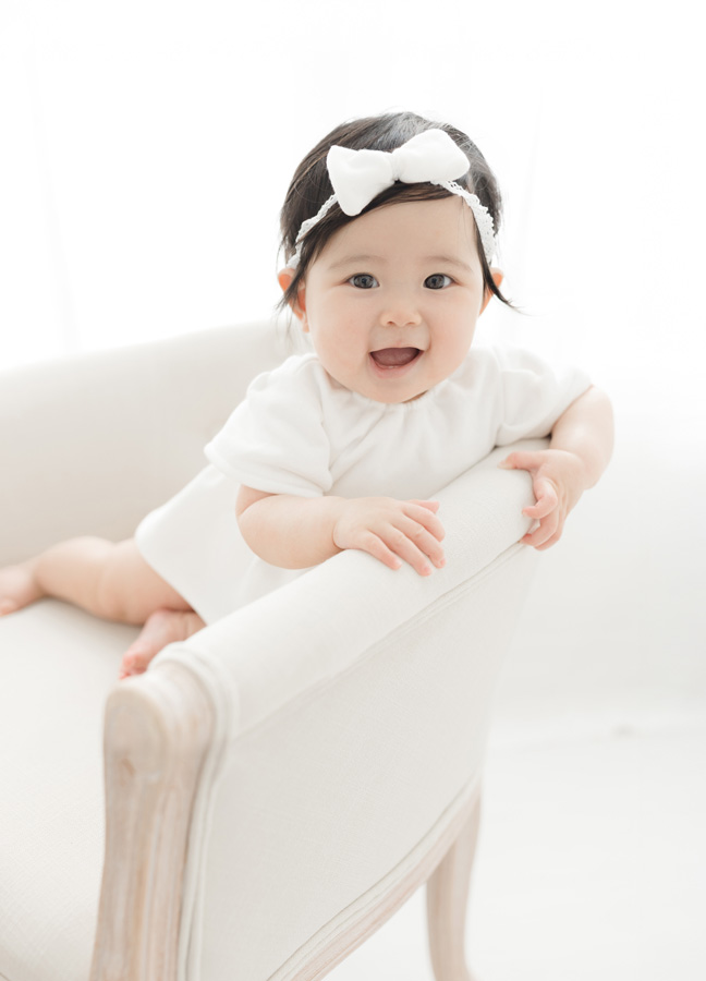 Smiling baby girl in a white outfit and bow headband posing in a bright studio, a cheerful example of newborn photos ideas Northern Virginia.