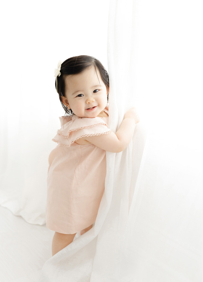 Smiling baby girl in a soft blush dress peeking around a white curtain, a playful example of newborn photos ideas Northern Virginia.