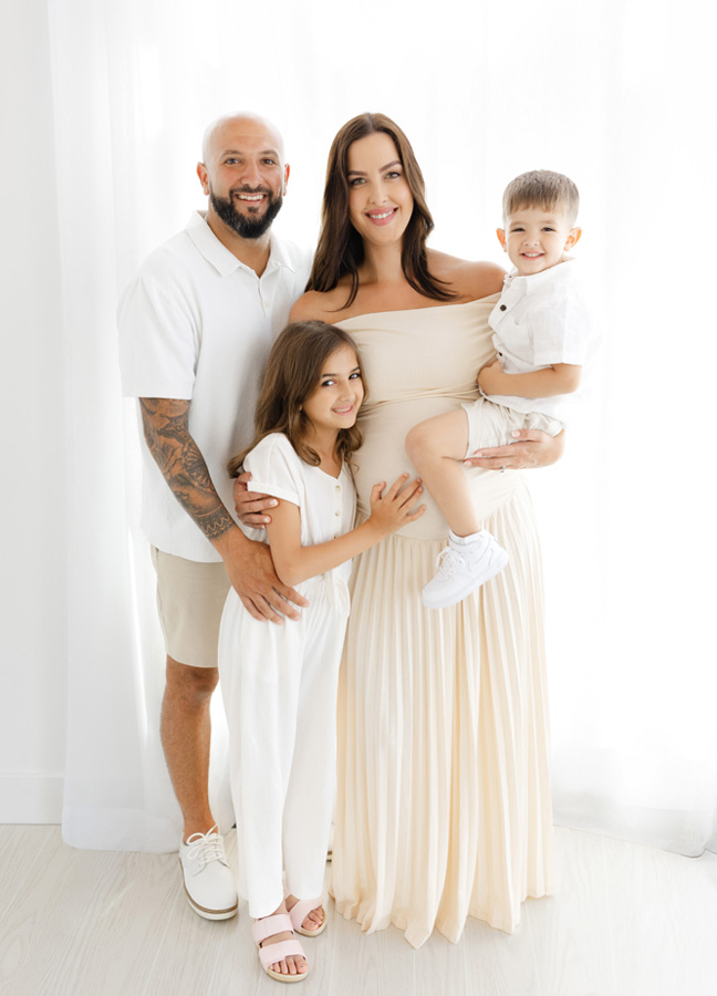 Family of four smiling together during studio maternity photos in Northern Virginia, with expecting mother in an ivory dress.